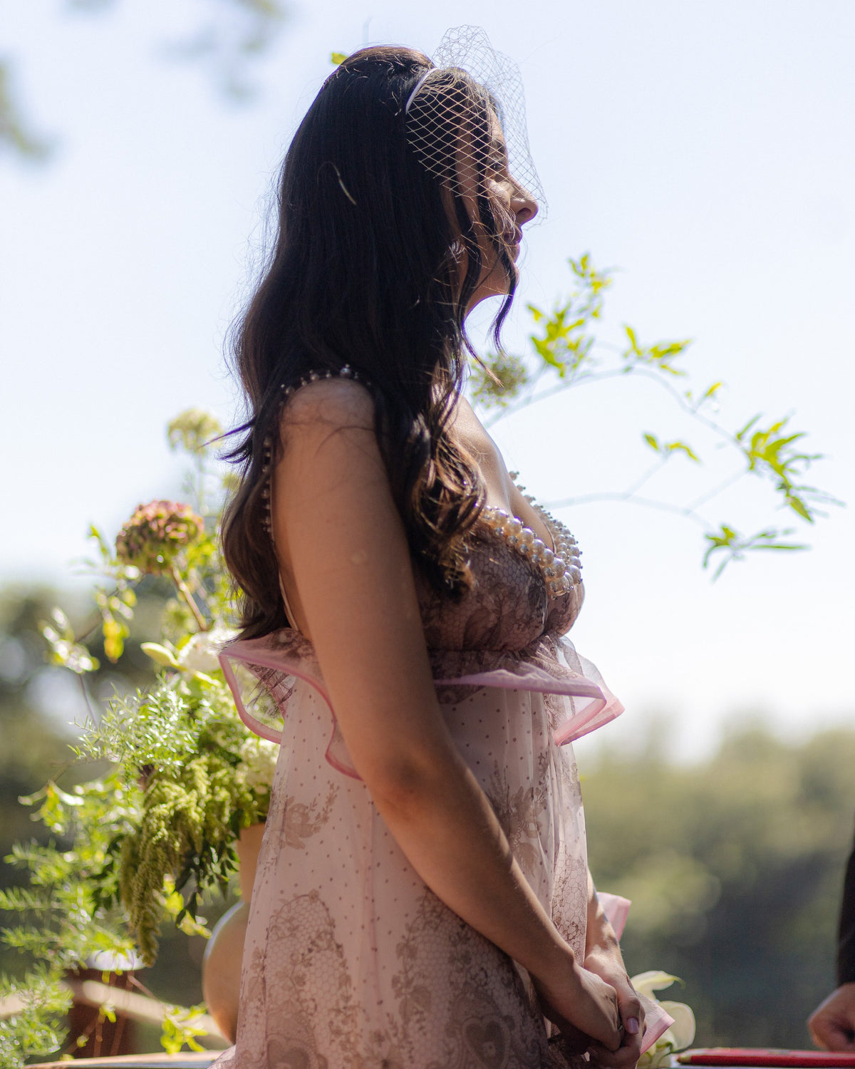 Caroline Duó de perfil, com vestido rosa, véu e mãos entrelaçadas.